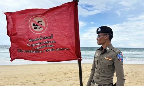 เจ้าหน้าที่ตำรวจท่องเที่ยวยืนข้าง “ธงแดง” สัญลักษณ์ “ห้ามเล่นน้ำ” ที่ปักไว้บริเวณชายหาดกะรน ภาพ: ตำรวจท่องเที่ยวภูเก็ต
